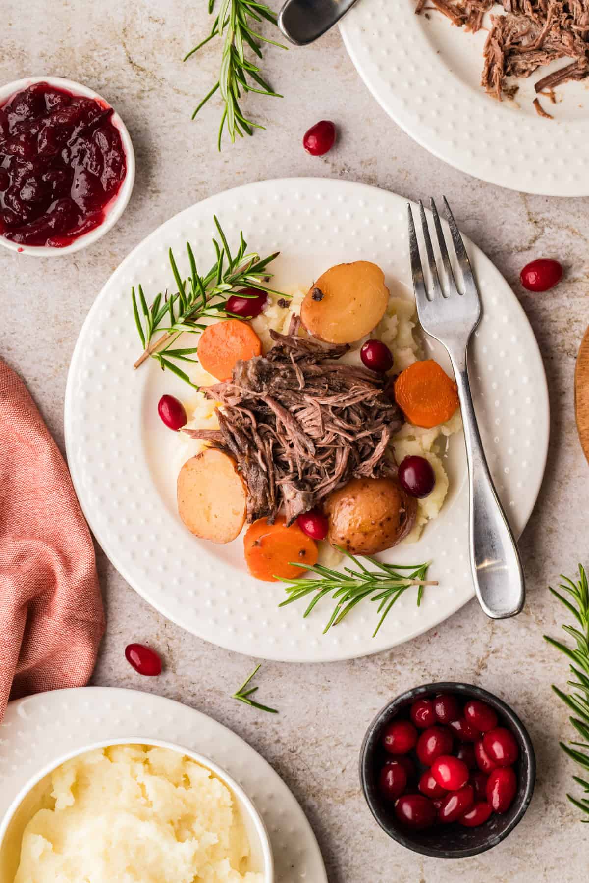 A plate with shredded beef, potatoes, carrots, and rosemary on mashed potatoes, surrounded by bowls of cranberries and cranberry sauce.