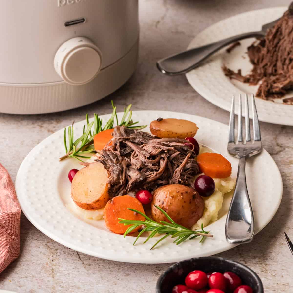 A plate with shredded cranberry beef, potatoes, carrots, and cranberries on mashed potatoes, garnished with rosemary, beside a fork and a slow cooker.