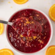 A spoonful of cranberry sauce garnished with orange zest held above a bowl, surrounded by orange slices on a white surface.