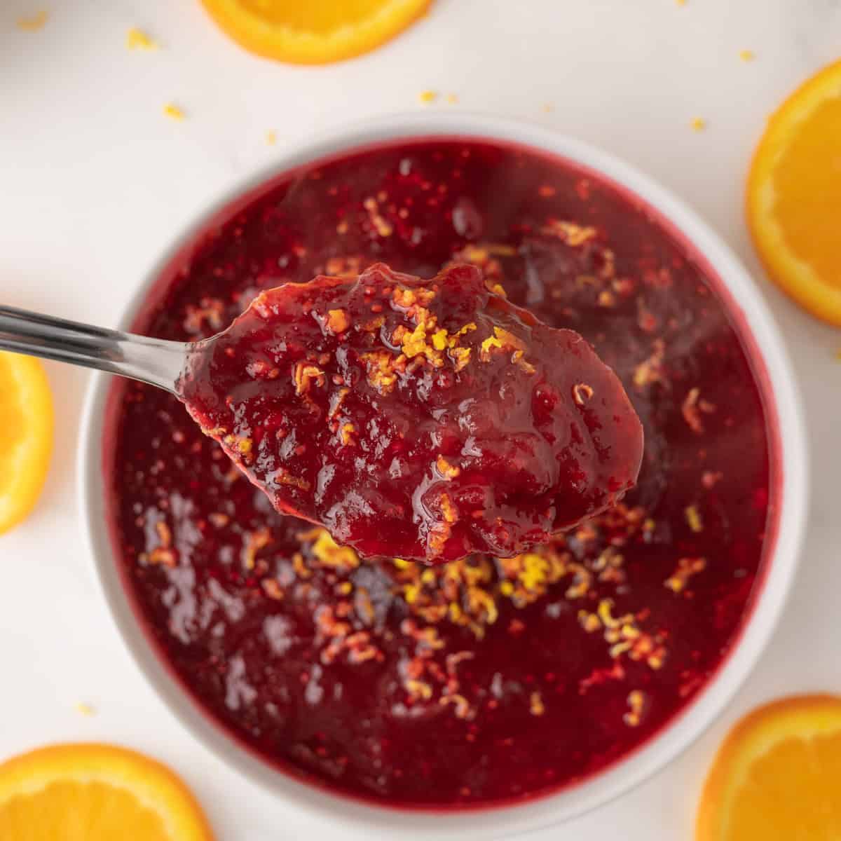 A spoonful of cranberry sauce garnished with orange zest held above a bowl, surrounded by orange slices on a white surface.
