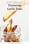 Close-up of Parmesan garlic fries in a glass jar, with one fry being held above the jar. Text on the image reads Parmesan Garlic Fries and by aweekendcook.com.