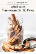 A mason jar filled with golden brown Parmesan garlic fries sits on a plate with a small metal dish of dipping sauce on the side.