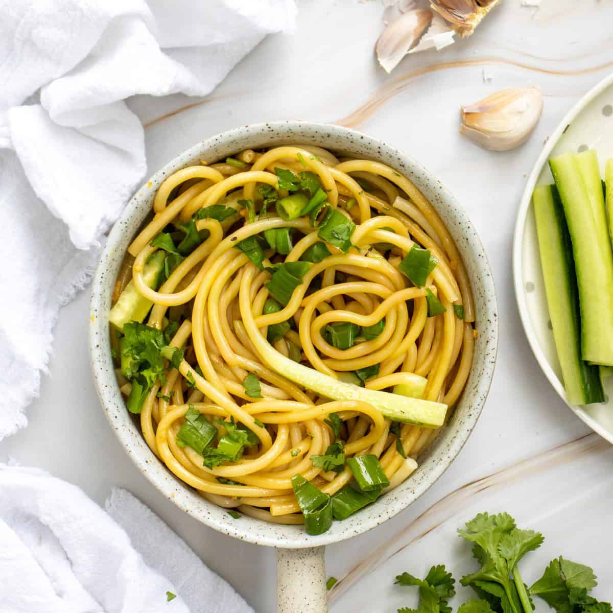 A bowl of noodles garnished with chopped green herbs and cucumber sticks, placed on a white surface with garlic, cucumber slices, and cilantro nearby.