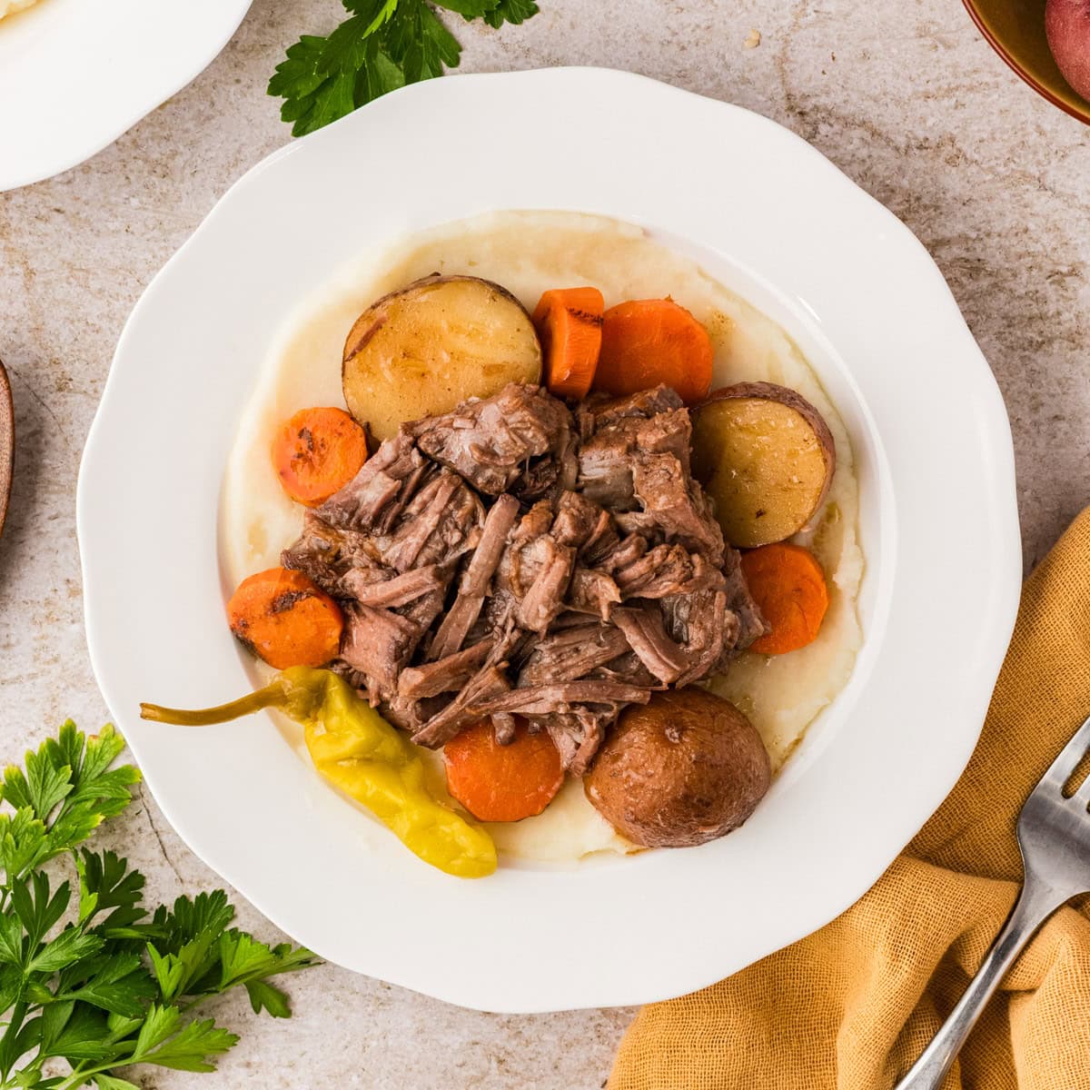 A plate of pot roast with shredded beef, carrots, potatoes, and a pepper served over mashed potatoes on a white plate.