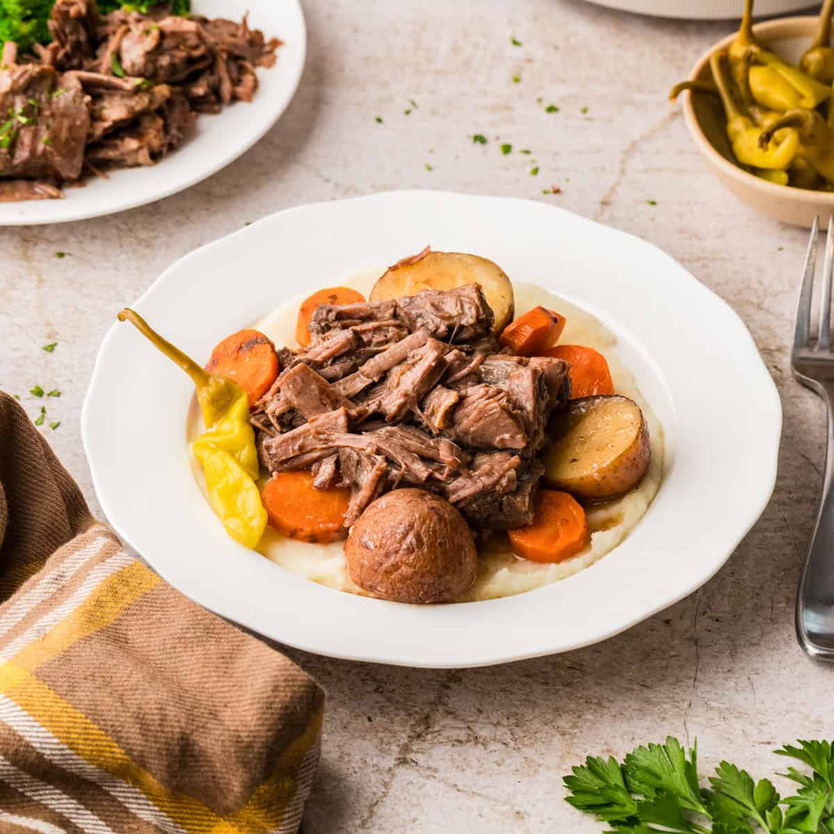 A plate of pot roast with shredded beef, potatoes, carrots, and pepperoncini served over mashed potatoes.