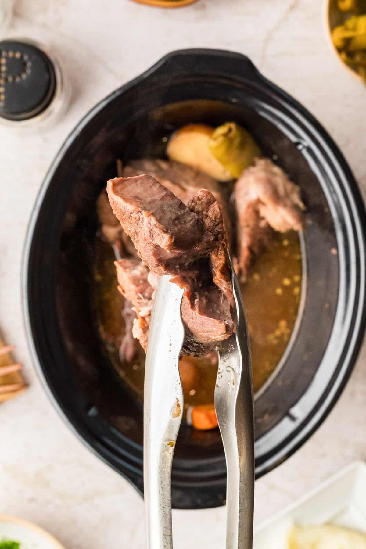 A pair of metal tongs holds a piece of cooked beef above a slow cooker filled with broth and vegetables.