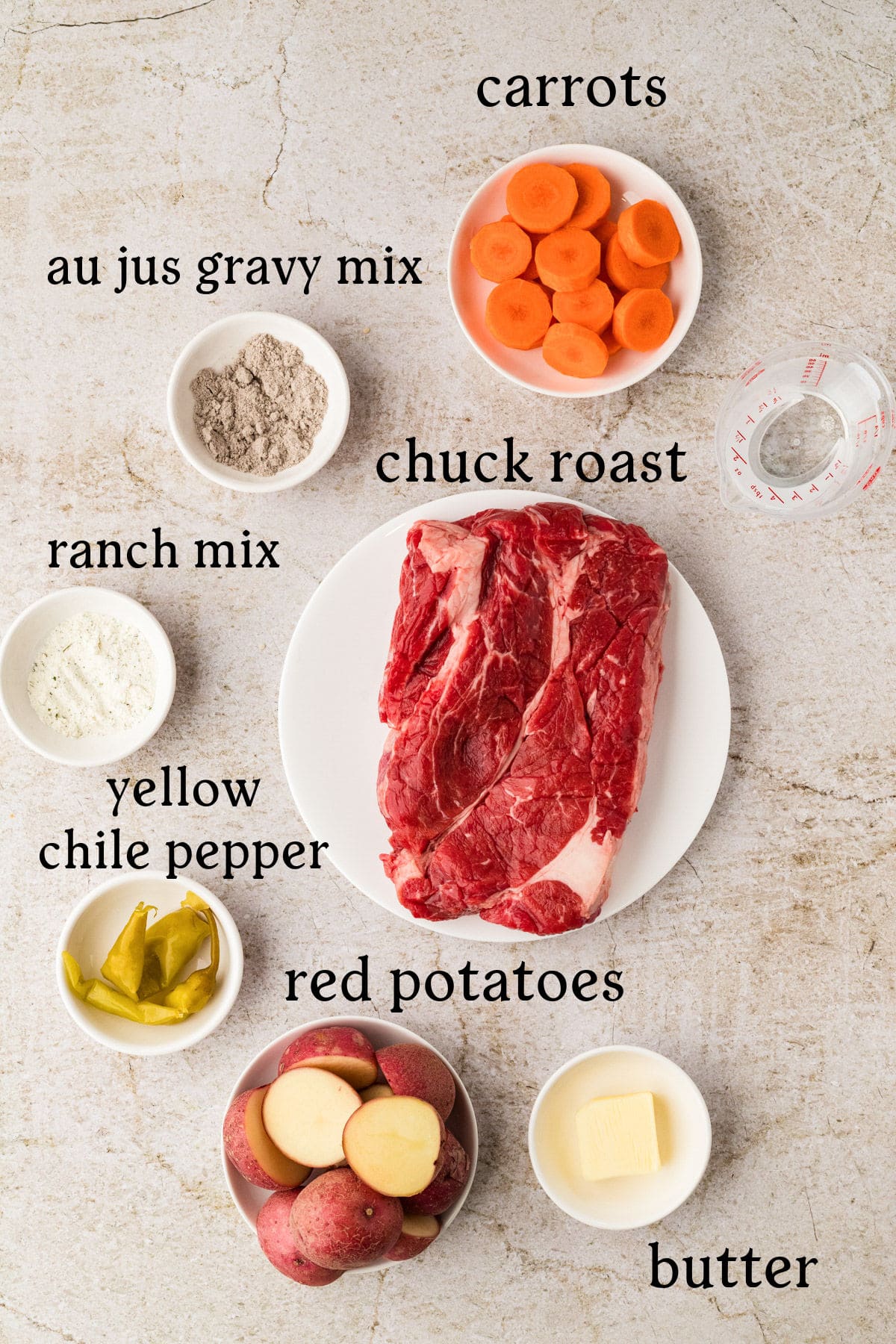 Top-down view of labeled ingredients for a chuck roast recipe, including chuck roast, carrots, red potatoes, butter, yellow chile pepper, ranch mix, au jus gravy mix, and water.