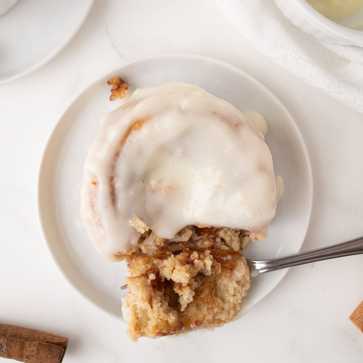 A cinnamon roll covered in white icing sits on a white plate, with a fork cutting into a bite-sized piece.