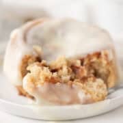 A close-up of a cinnamon roll with white icing, partially cut with a fork on a white plate.