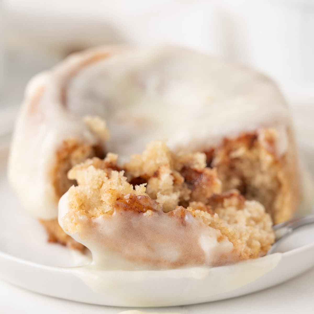 A close-up of a cinnamon roll with white icing, partially cut with a fork on a white plate.