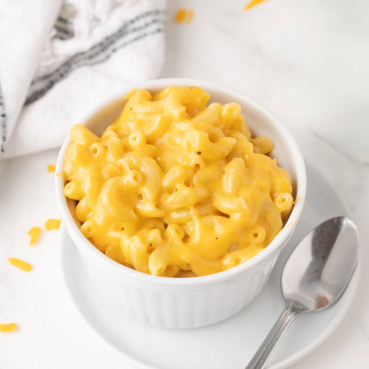 A white ramekin filled with creamy macaroni and cheese sits on a white plate with a metal spoon and a cloth napkin nearby.
