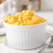 A white ramekin filled with creamy macaroni and cheese sits on a saucer next to a spoon, with a blurred background.