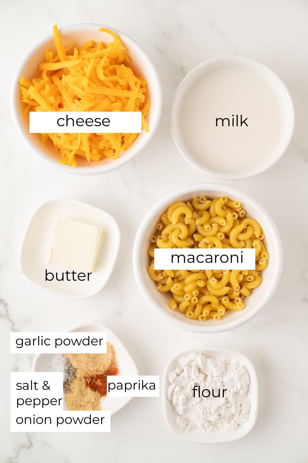 Bowls containing shredded cheese, milk, butter, uncooked macaroni, flour, and a mix of salt, pepper, paprika, and garlic powder arranged on a white surface.