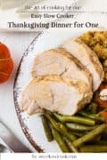 A plate with two slices of turkey breast, green beans, stuffing, and carrots, set on a white table with a decorative orange pumpkin nearby.