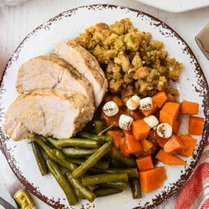 A plate with sliced turkey, green beans, stuffing, and sweet potatoes topped with mini marshmallows on a white wooden table.