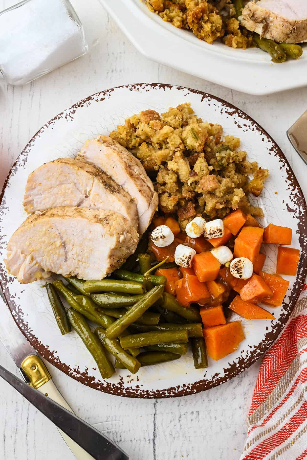 A plate with sliced turkey, green beans, stuffing, and sweet potatoes topped with mini marshmallows on a white wooden table.