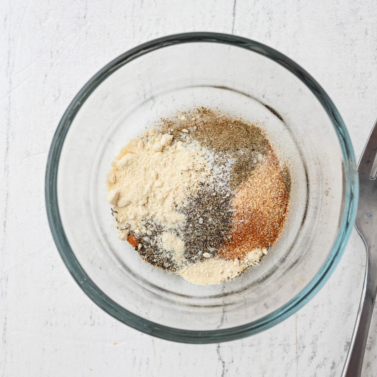 A glass bowl containing assorted dry spices and seasonings, seen from above, on a white surface next to a metal fork.
