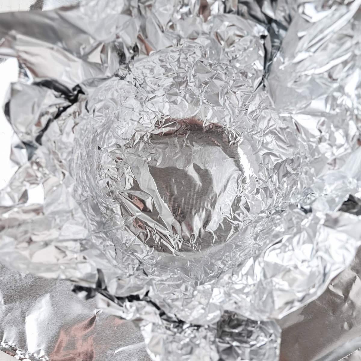 A close-up view of crumpled and shiny aluminum foil forming a bowl or cup shape, reflecting light and creating various textures.