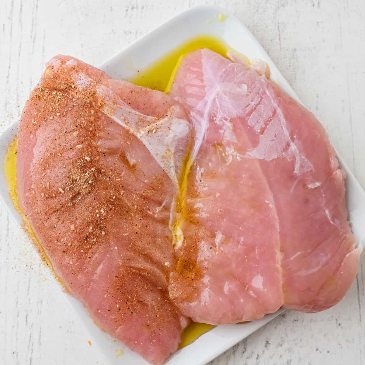 Two raw chicken breasts on a white plate, one seasoned with spices and oil, the other plain—perfect for preparing a simple Thanksgiving dinner for one on a light-colored surface.