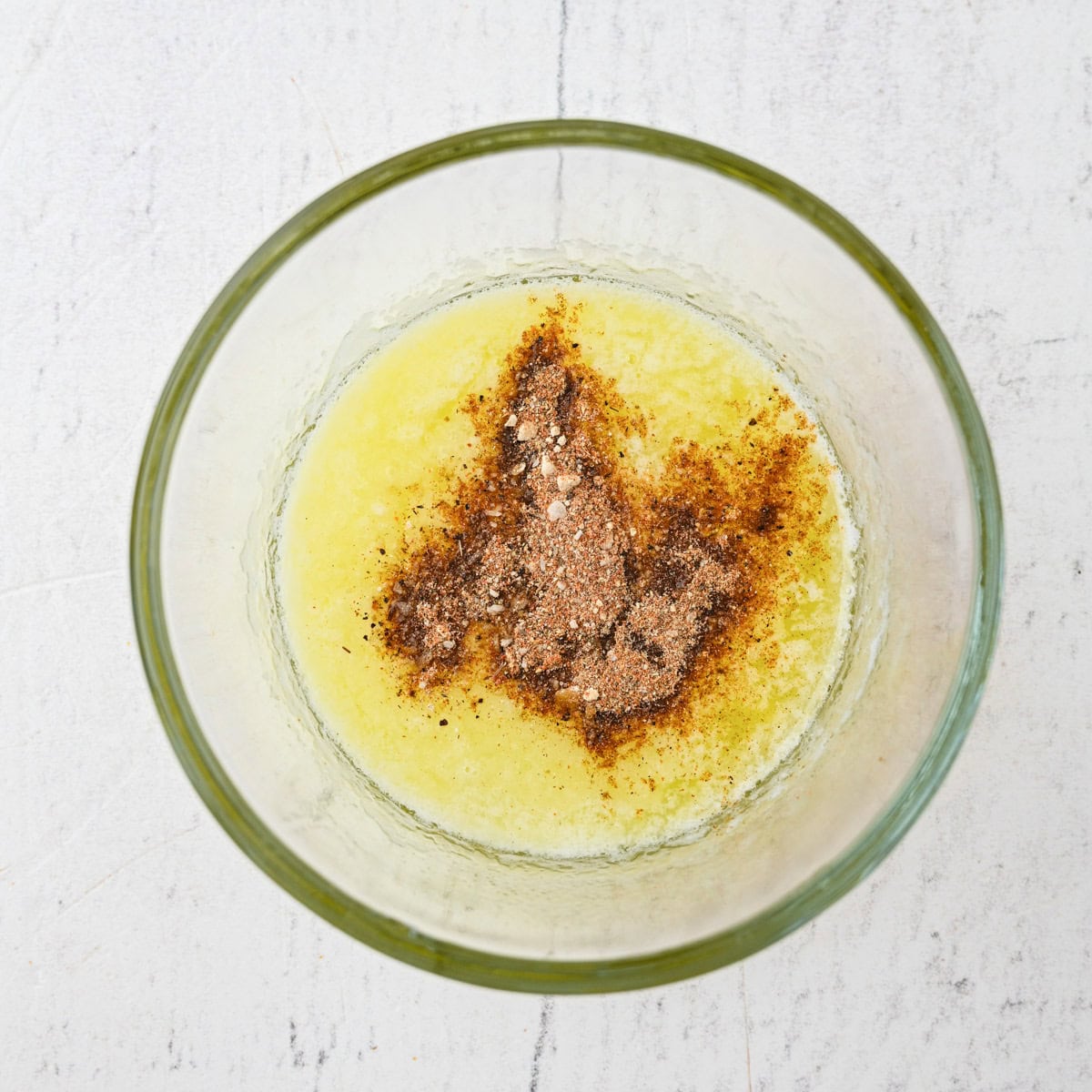 A glass bowl containing melted butter topped with a mix of ground spices on a white surface.