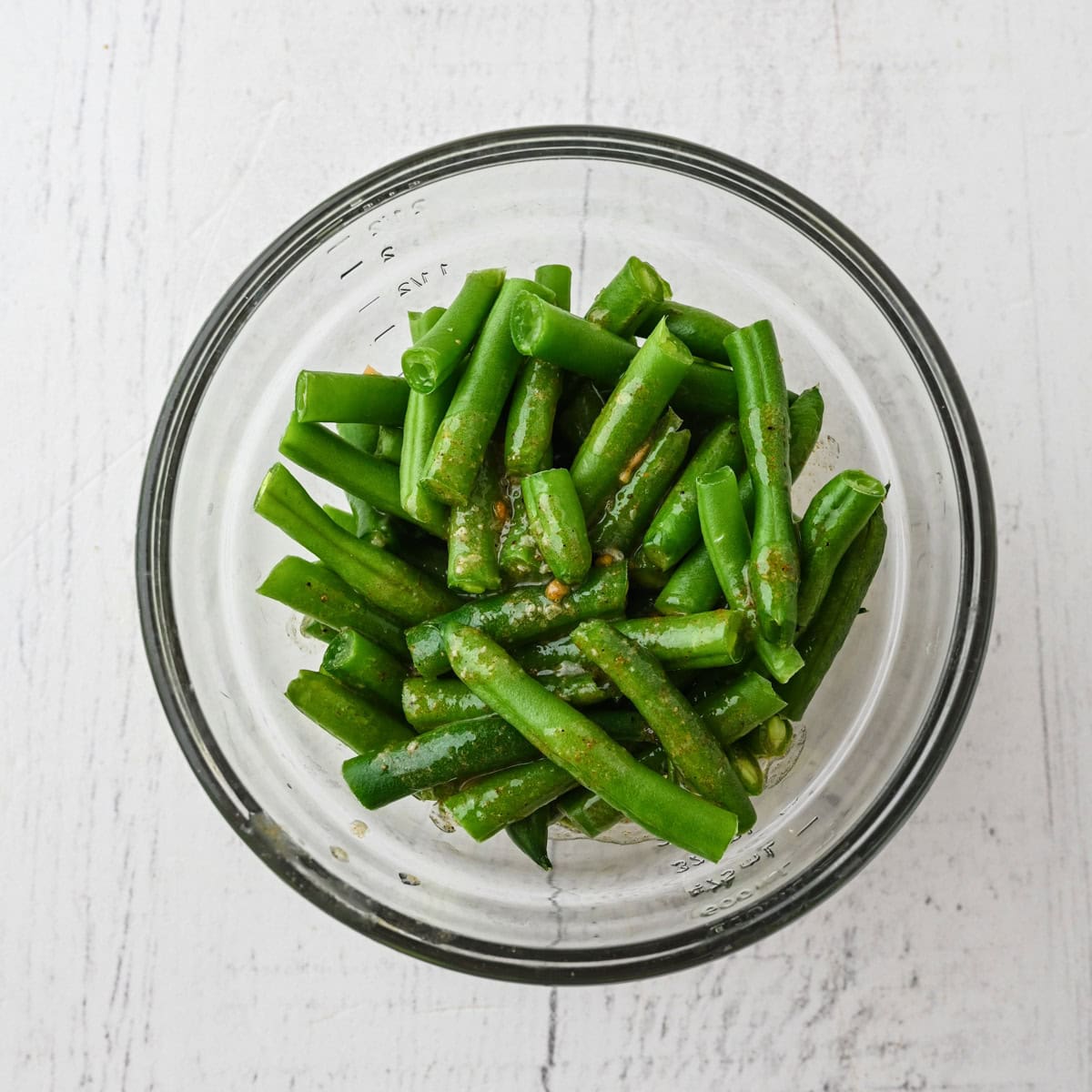 A glass bowl containing cut green beans with a light coating of dressing, perfect for a simple Thanksgiving dinner for one, placed on a white wooden surface.