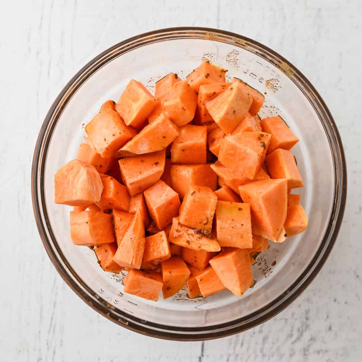 A glass bowl filled with cubed sweet potatoes seasoned with spices, perfect for a cozy Thanksgiving dinner for one, placed on a white surface.