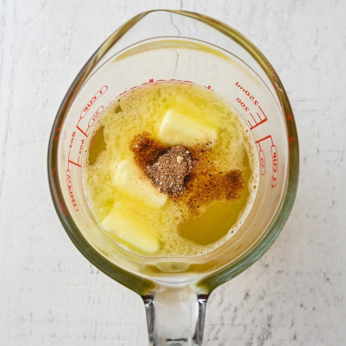A glass measuring cup with melted butter, pats of unmelted butter, and a heap of ground spices sits ready on a white surface—perfect for preparing a cozy Thanksgiving dinner for one.