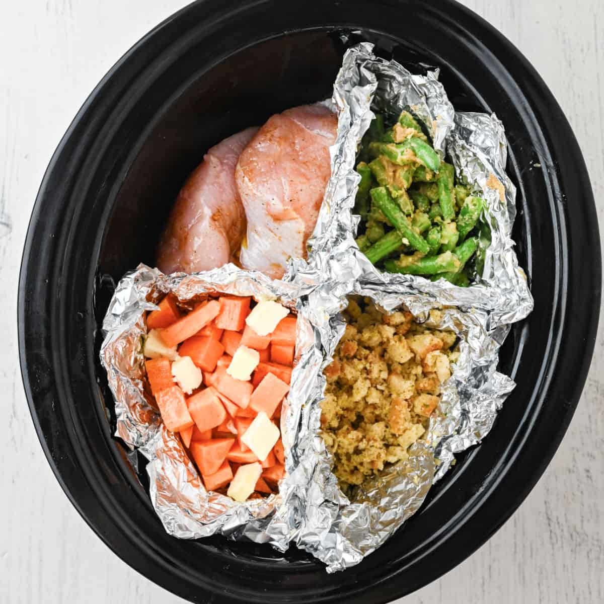 A slow cooker with foil dividers holds raw chicken breast, green beans, sweet potatoes, and stuffing—each ingredient uncooked and sectioned separately for an easy Thanksgiving dinner for one.