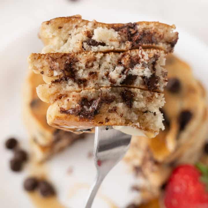 A fork holds a sliced stack of chocolate chip pancakes, with a blurred plate, chocolate chips, and a strawberry in the background.
