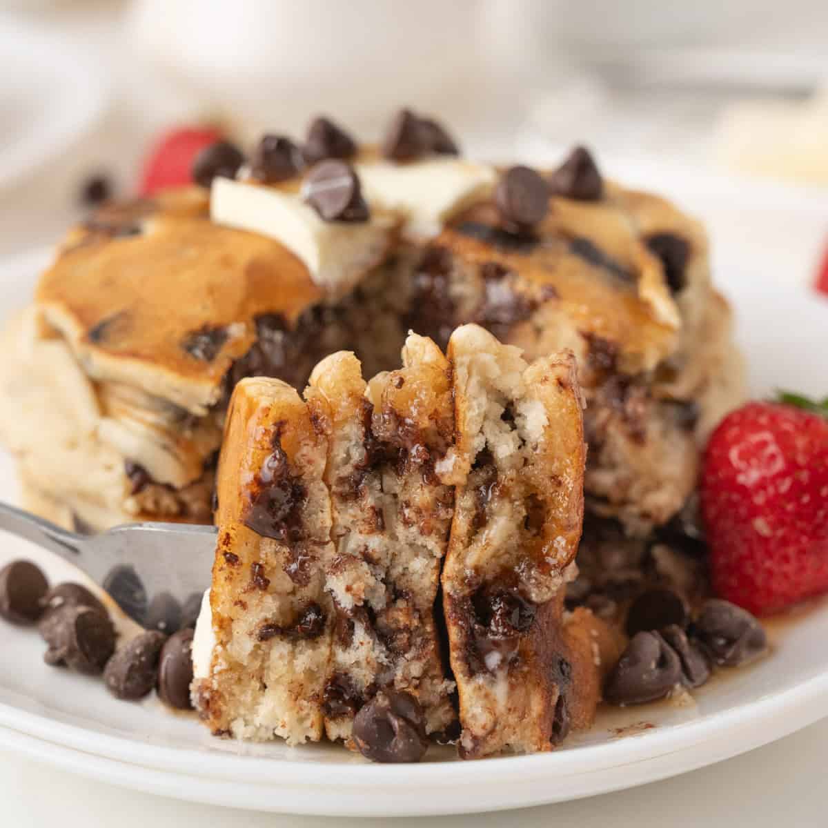 A close-up of chocolate chip pancakes with melted chocolate chips, a pat of butter on top, and a fork holding a bite. Strawberries and extra chocolate chips are on the plate.