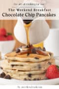 A stack of chocolate chip pancakes is topped with chocolate chips and being drizzled with syrup, with strawberries on the side.