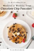 A plate of chocolate chip pancakes topped with butter and syrup, surrounded by strawberries and chocolate chips, with a fork and extra pancakes nearby.