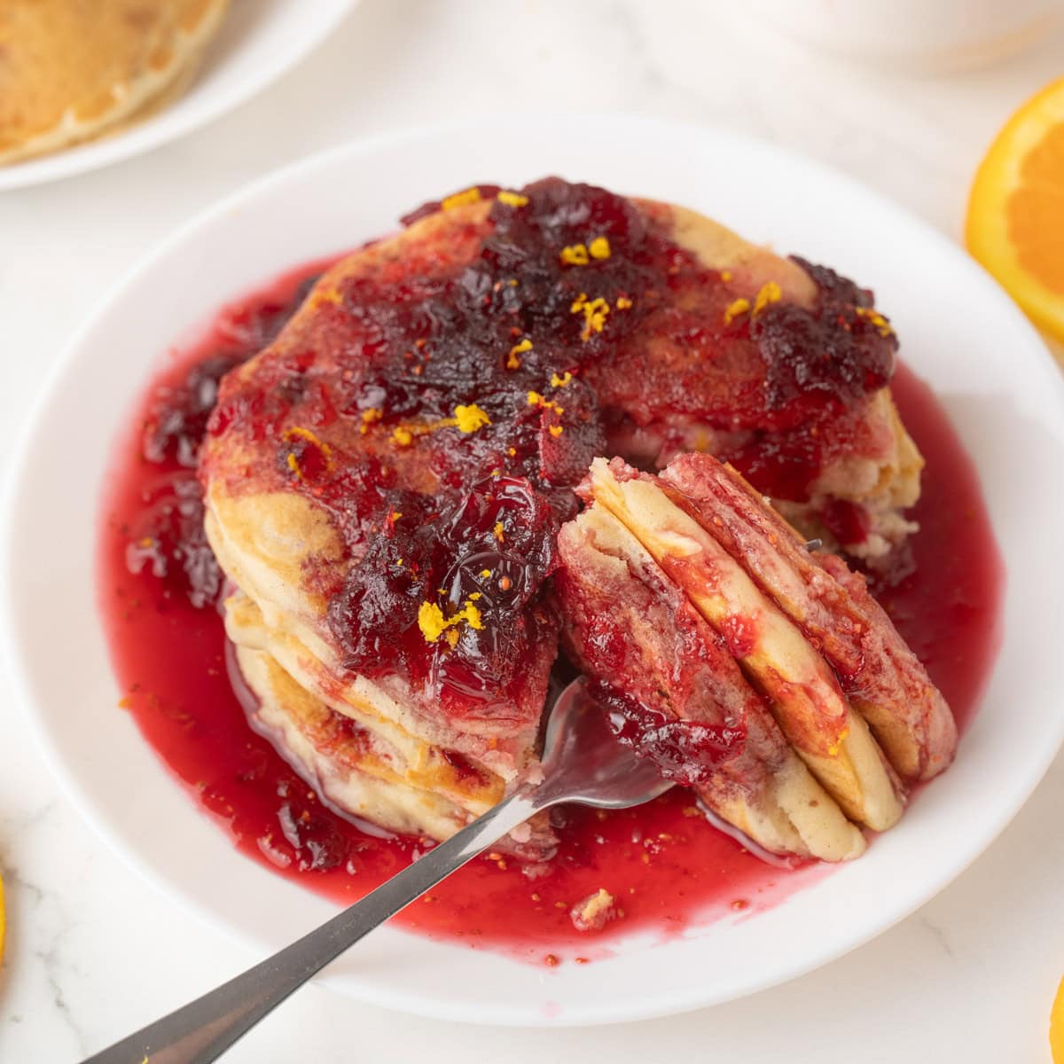 A stack of cranberry orange pancakes topped with cranberry syrup and orange zest on a white plate, with a fork cutting into the pancakes.