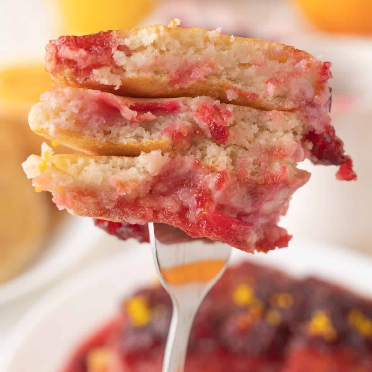A close-up of a fork holding a stack of cranberry orange pancakes with a cranberry syrup, showing the soft, fluffy texture and vibrant red fruit topping.