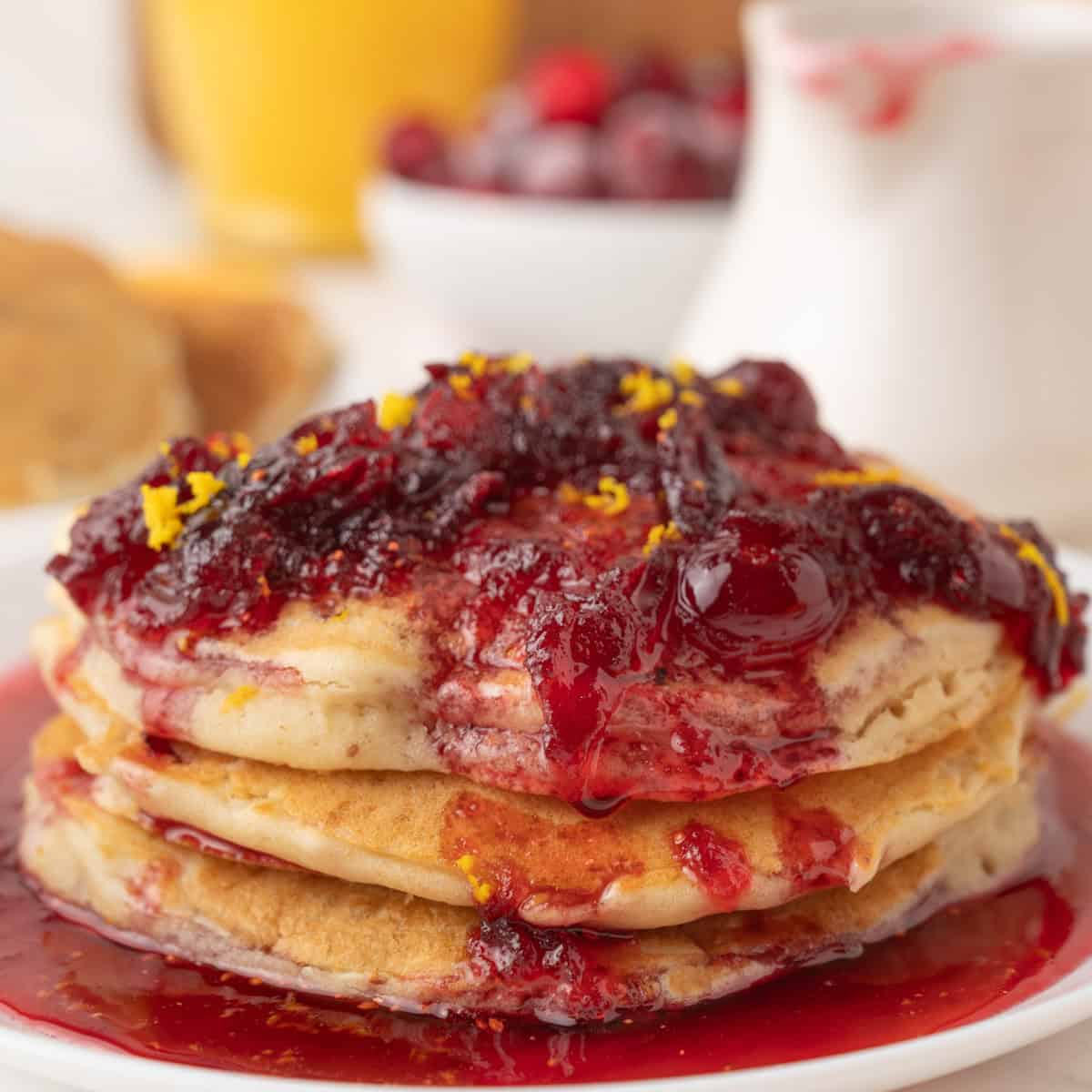 A stack of cranberry pancakes topped with thick cranberry syrup and orange zest sits on a white plate, with a blurred bowl of cherries and glass of orange juice in the background.