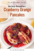 A stack of cranberry pancakes topped with cranberry orange sauce, partially sliced, served on a white plate with orange slices in the background. Perfect pancakes for one craving a burst of citrus and berry flavor.