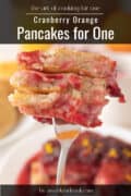 A close-up of a fork holding a bite of cranberry pancakes, with a plate of pancakes blurred in the background. Text on the image reads Cranberry Orange Pancakes for One.
