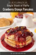 A stack of cranberry orange pancakes, perfect as pancakes for one, topped with cranberry sauce and orange zest, served on a white plate with syrup and extra pancakes in the background.