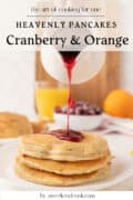 A stack of cranberry pancakes on a plate is drizzled with vibrant cranberry syrup; orange, cranberries, juice, and another pancake stack add a fresh touch in the background.