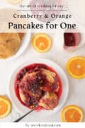 A plate of cranberry pancakes and orange pancakes topped with cranberry sauce, surrounded by fresh orange slices, with a fork and extra pancakes for one on the side.