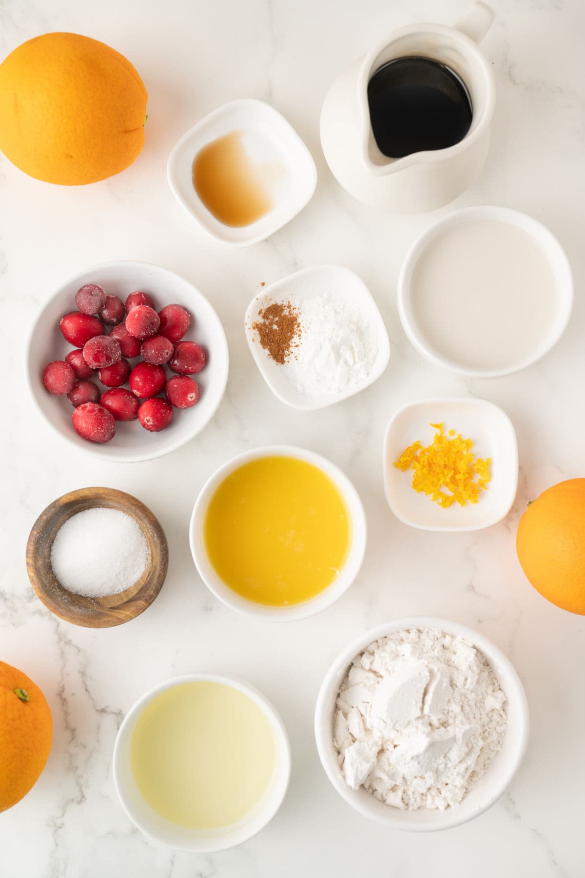 Assorted baking ingredients including flour, sugar, cranberries, orange zest, spices, vanilla, melted butter, oil, milk, syrup, and whole oranges arranged on a white surface.