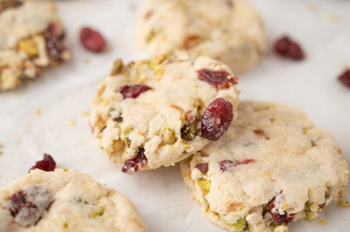 Three crumbly cookies with visible pieces of dried cranberries and chopped pistachios on white parchment paper.