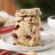 A stack of four thick cookies with visible pieces of dried cranberries and pistachios sits on a white plate.