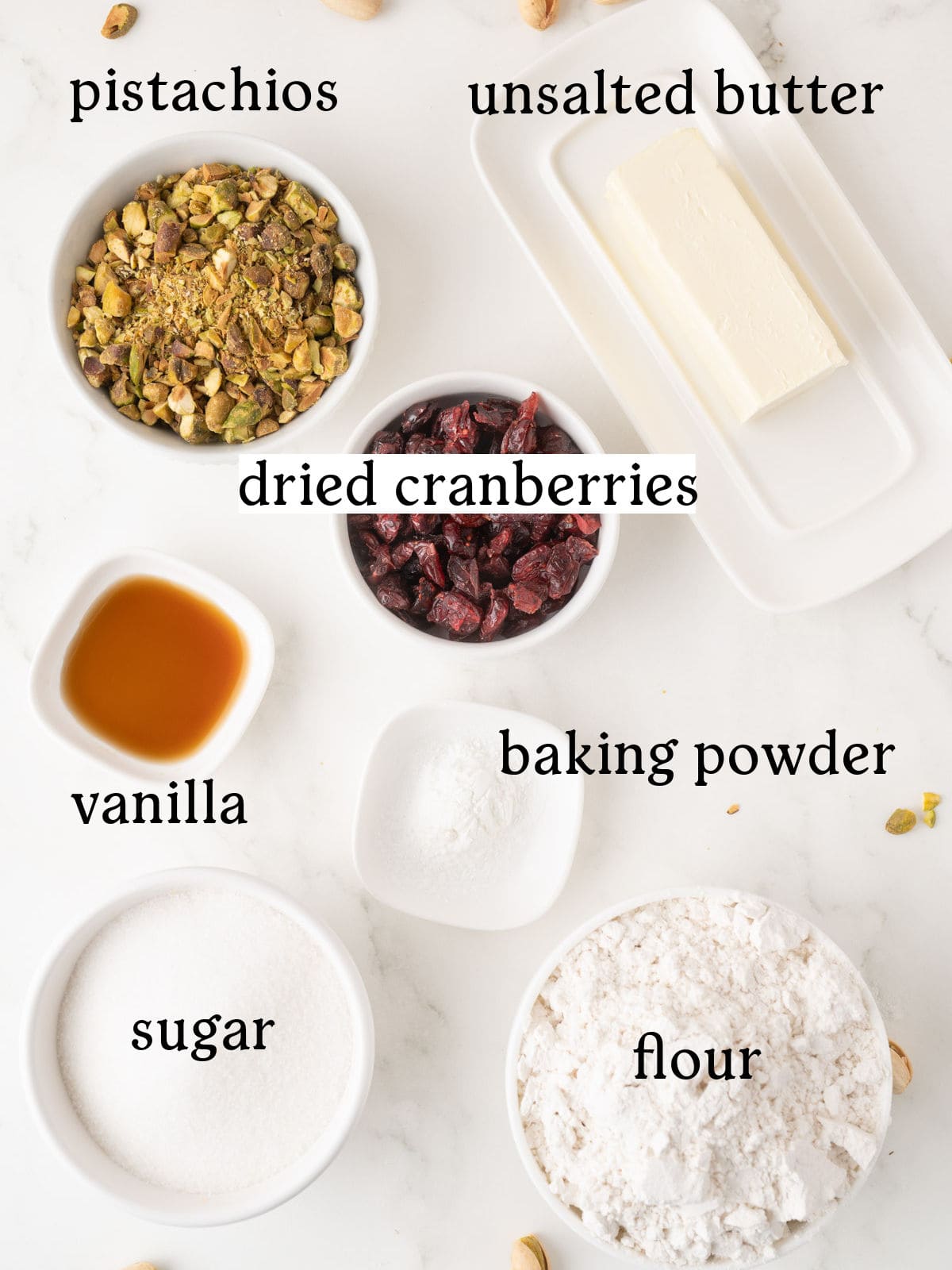 Various baking ingredients including pistachios, dried cranberries, unsalted butter, vanilla, baking powder, sugar, and flour are arranged in bowls on a white surface.