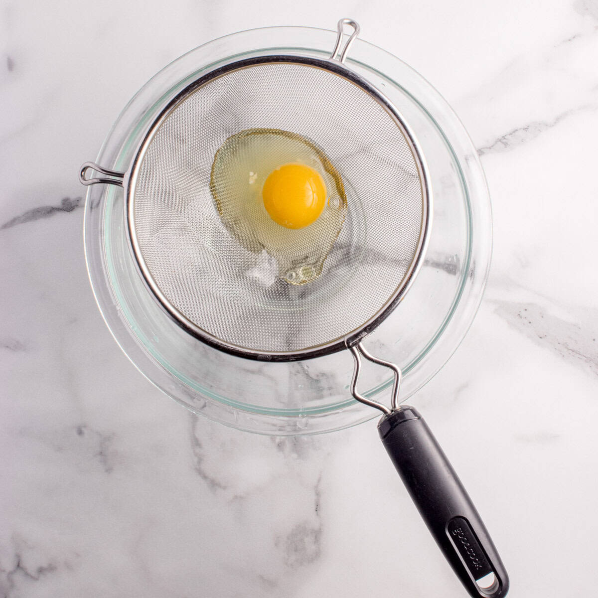 A raw egg is placed in a fine mesh strainer over a clear glass bowl on a white marble surface.