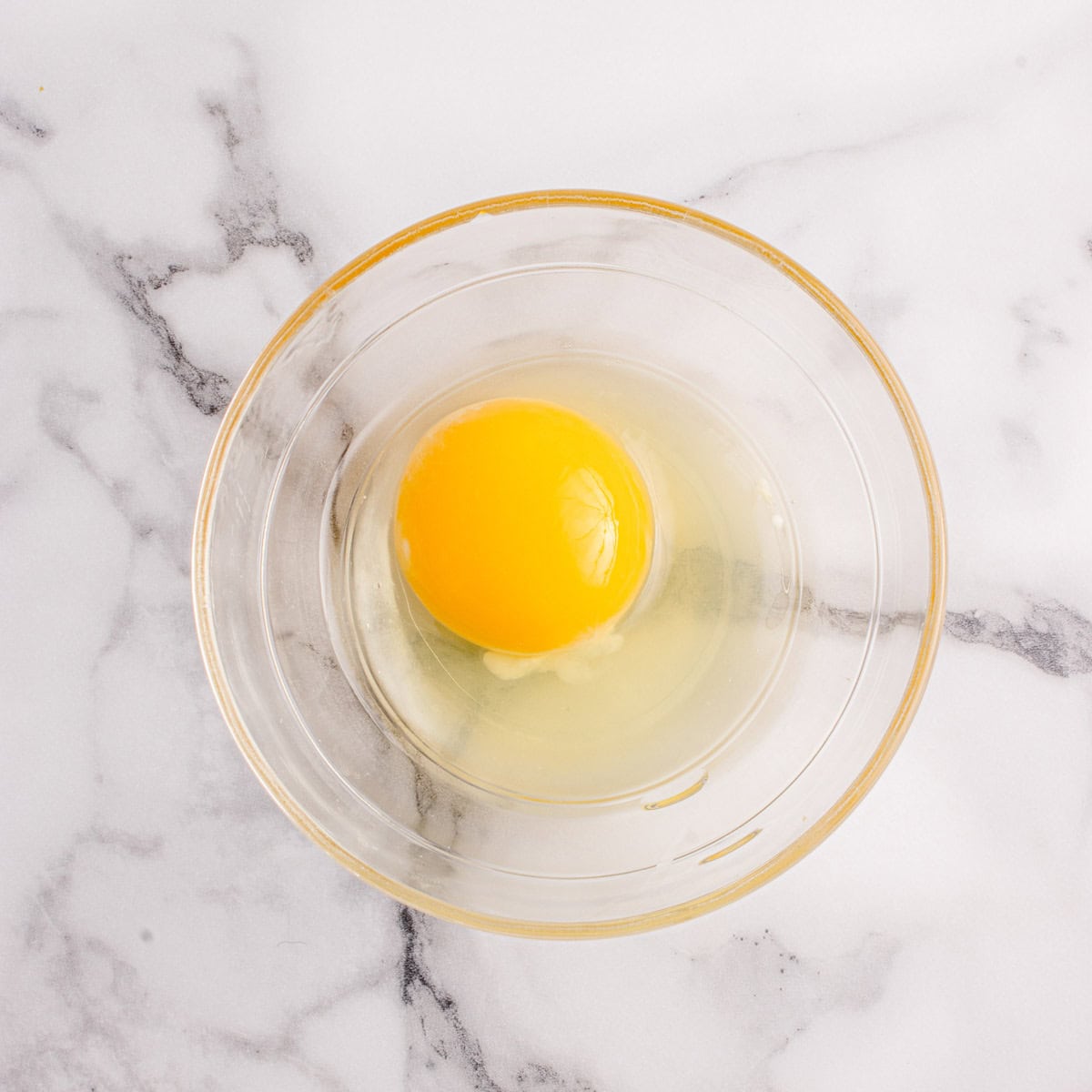 A raw egg with an intact yolk and clear egg white in a glass bowl on a white marble surface.