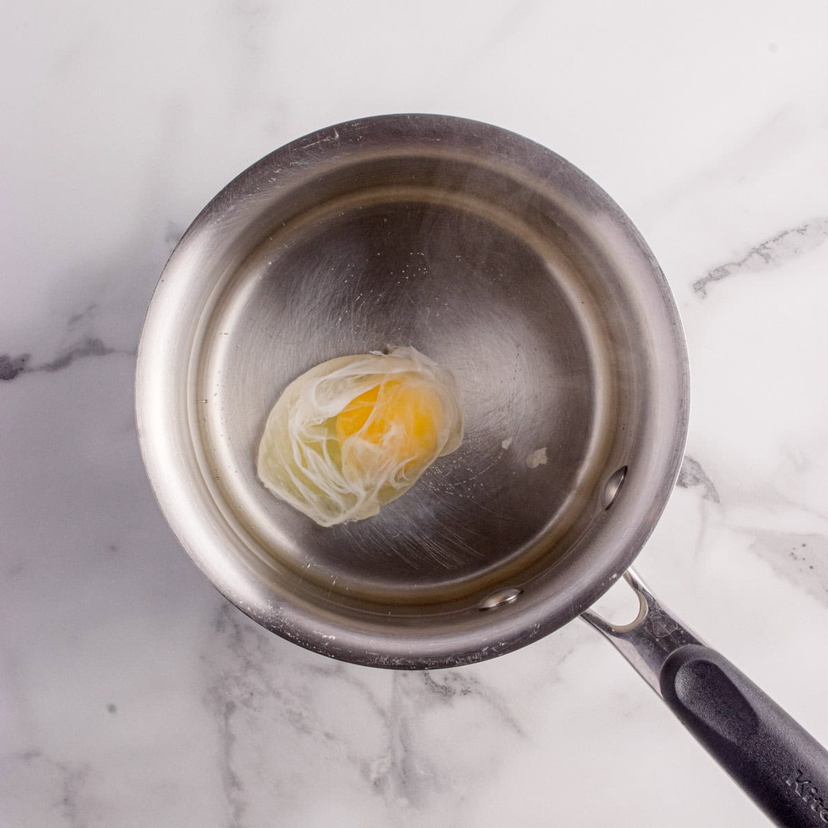 A poached egg sits in a small stainless steel pot on a marble surface.