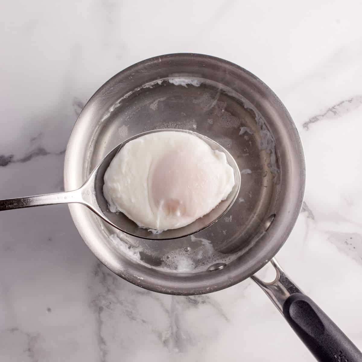 A slotted spoon holding a poached egg above a small saucepan filled with water on a white marble surface.