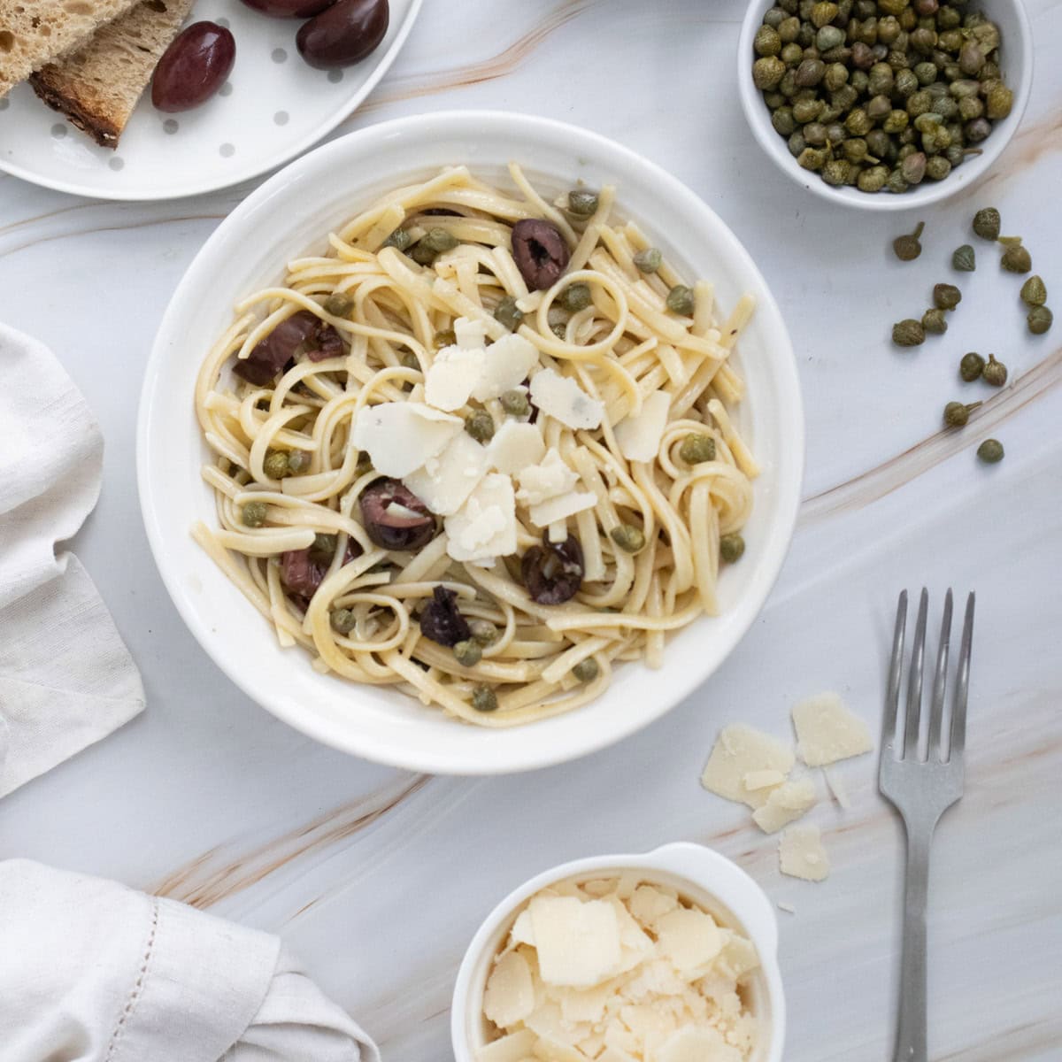 A plate of linguine pasta with shaved cheese, capers, and olives on a white table, next to a fork, a bowl of shaved cheese, and bread.