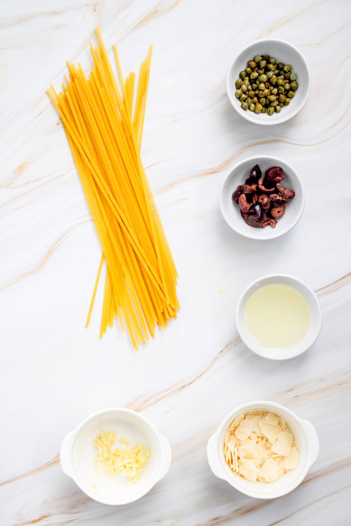 Uncooked pasta, capers, sliced olives, lemon juice, minced garlic, and grated cheese are arranged in small bowls on a white marble surface.